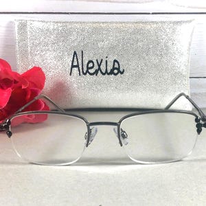 May include: A pair of rimless eyeglasses with dark metal frames and decorative black accents. A silver case with the name "Alexis" in black script is behind the glasses. A red flower is to the left of the glasses.