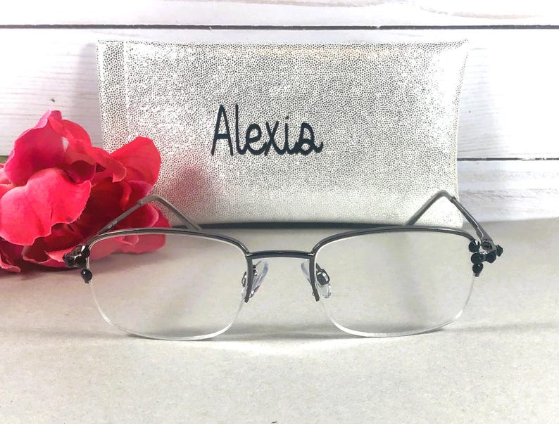 May include: A pair of rimless eyeglasses with dark metal frames and decorative black accents. A silver case with the name "Alexis" in black script is behind the glasses. A red flower is to the left of the glasses.