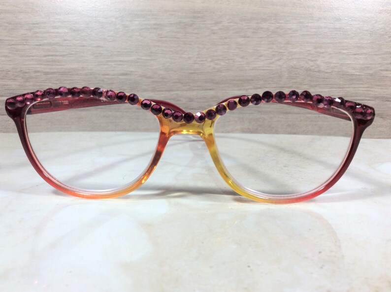 May include: Eyeglasses with a gradient frame, transitioning from red to yellow. The top of the frame is adorned with a row of purple rhinestones. The glasses have a cat-eye shape, suitable for various face shapes.