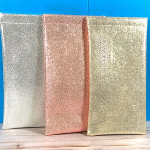Op de afbeelding: Drie rechthoekige etuis met een glinsterende, textuurrijke afwerking. Het linker etui is zilverkleurig, het middelste etui is ros&eacute;goud en het rechter etui is goudkleurig. Elk etui heeft een gladde, platte bovenrand en een verticale vorm.