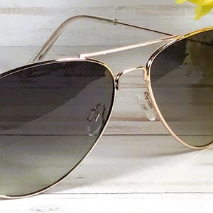 May include: Aviator sunglasses with a gold frame and gradient lenses. The lenses transition from dark to light. The sunglasses are set against a white wooden background with yellow flowers.