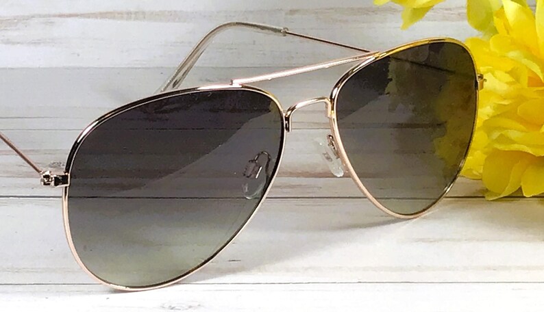 May include: Aviator sunglasses with a gold frame and gradient lenses. The lenses transition from dark to light. The sunglasses are set against a white wooden background with yellow flowers.