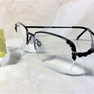 May include: Black rimless spectacles with clear lenses. The frames have a decorative black embellishment on the outer edges. The temples are thin and black. The spectacles are resting on a light-coloured surface.