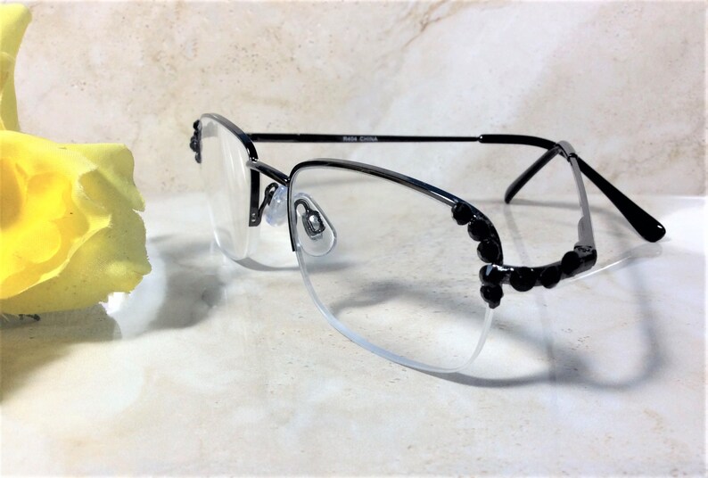 May include: Black rimless spectacles with clear lenses. The frames have a decorative black embellishment on the outer edges. The temples are thin and black. The spectacles are resting on a light-coloured surface.