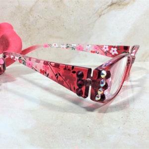 Puede incluir: Gafas rojas con un estampado floral en las monturas y varillas. Las monturas est&aacute;n adornadas con peque&ntilde;os brillantes iridiscentes. Las lentes son transparentes. Las gafas se presentan sobre un fondo suave y neutro.