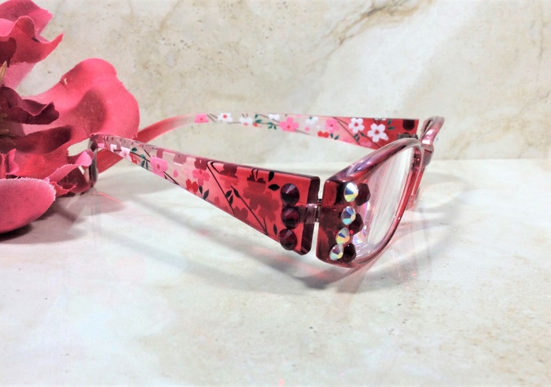 Puede incluir: Gafas rojas con un estampado floral en las monturas y varillas. Las monturas est&aacute;n adornadas con peque&ntilde;os brillantes iridiscentes. Las lentes son transparentes. Las gafas se presentan sobre un fondo suave y neutro.