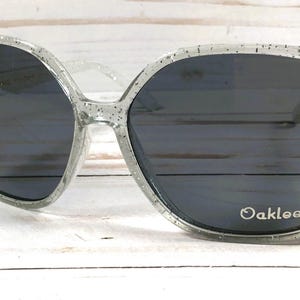 May include: Clear glitter sunglasses with black lenses and the word "Oaklee" printed on the right lens.