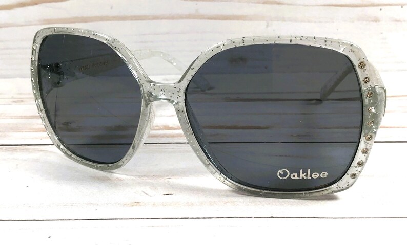 May include: Clear glitter sunglasses with black lenses and the word "Oaklee" printed on the right lens.