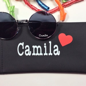 以下が含まれることがあります： 黒色の眼鏡ケースに「Camila」という名前が白で印刷され、赤いハートが描かれています。ケースは閉じており、その上に黒い丸いサングラスが置かれています。