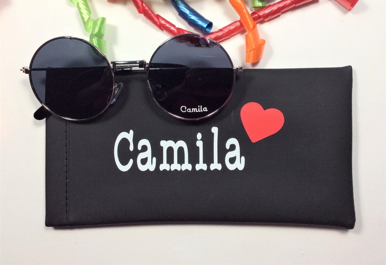 以下が含まれることがあります： 黒色の眼鏡ケースに「Camila」という名前が白で印刷され、赤いハートが描かれています。ケースは閉じており、その上に黒い丸いサングラスが置かれています。