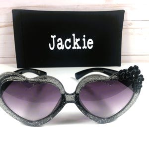 May include: Black heart-shaped sunglasses with glitter accents and black flower embellishments. The sunglasses come with a black case that has the name "Jackie" printed on it.