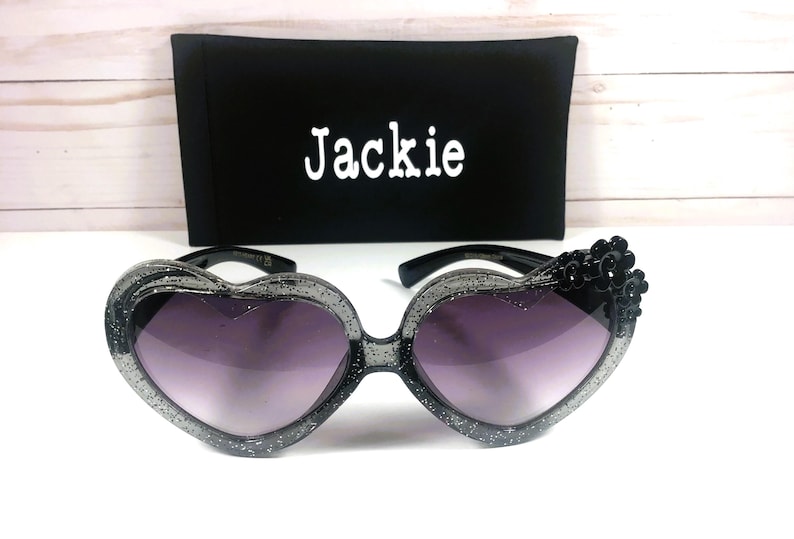 May include: Black heart-shaped sunglasses with glitter accents and black flower embellishments. The sunglasses come with a black case that has the name "Jackie" printed on it.