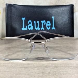 Op de afbeelding: Bril met zilveren montuur, heldere glazen en decoratieve iriserende strass-steentjes op de slapen. Een zwarte brillenkoker met de naam "Laurel" in lichtblauwe letters staat achter de bril. De bril ligt op een licht houten oppervlak.