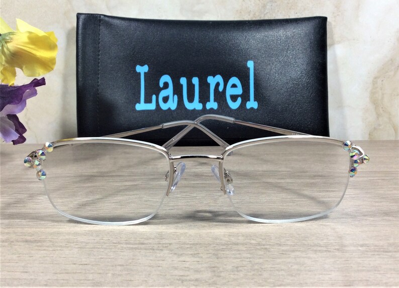 Op de afbeelding: Bril met zilveren montuur, heldere glazen en decoratieve iriserende strass-steentjes op de slapen. Een zwarte brillenkoker met de naam "Laurel" in lichtblauwe letters staat achter de bril. De bril ligt op een licht houten oppervlak.