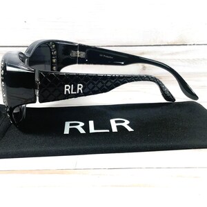 May include: Black sunglasses with a glossy finish and a decorative pattern on the temples. The brand name "RLR" is visible on the temples and the included black case. The sunglasses are resting on top of the case.