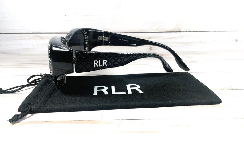 May include: Black sunglasses with a glossy finish and a decorative pattern on the temples. The brand name "RLR" is visible on the temples and the included black case. The sunglasses are resting on top of the case.