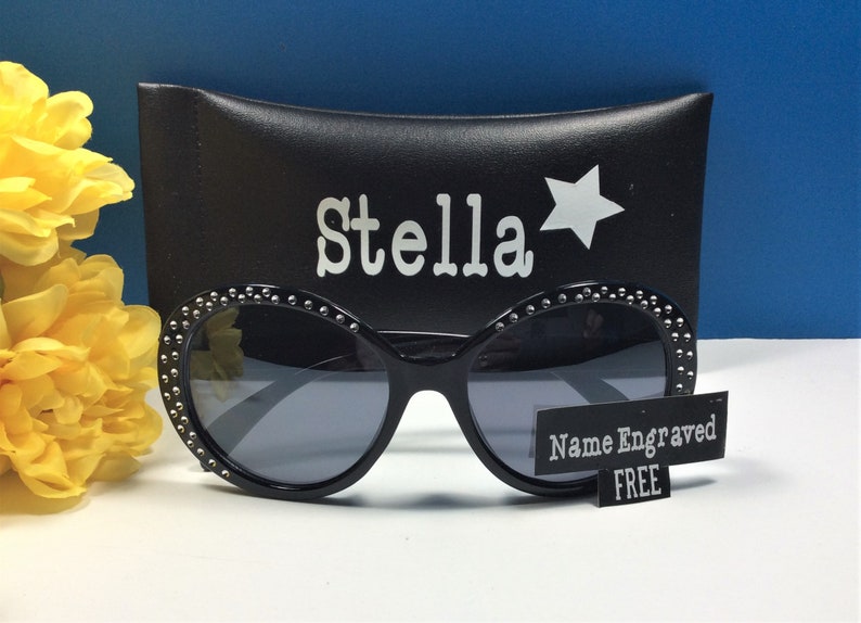 May include: Black sunglasses with a rhinestone trim and a black case with the name "Stella" and a star printed on it. The sunglasses come with a free name engraving.