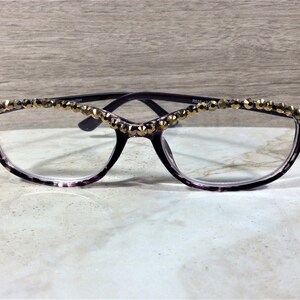 May include: Purple tortoiseshell spectacles featuring a row of faceted gold-tone studs along the top of the frames. The glasses have clear lenses and a cat-eye shape. The inner temple is marked with the text R825 42-16.