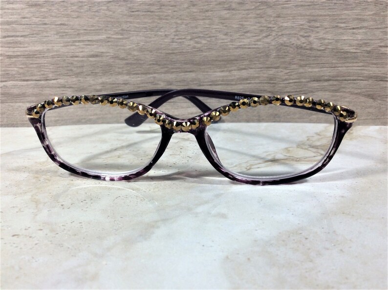 May include: Purple tortoiseshell spectacles featuring a row of faceted gold-tone studs along the top of the frames. The glasses have clear lenses and a cat-eye shape. The inner temple is marked with the text R825 42-16.