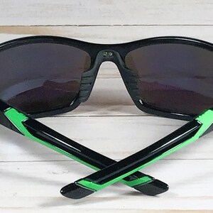 May include: Black sunglasses with dark lenses and green accents on the temples. The sunglasses are set against a light wood-grain background. The arms of the sunglasses are crossed.