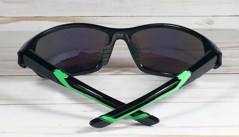May include: Black sunglasses with dark lenses and green accents on the temples. The sunglasses are set against a light wood-grain background. The arms of the sunglasses are crossed.