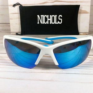 May include: White sports sunglasses with reflective blue lenses and bright blue temple tips. Behind the sunglasses is a black drawstring pouch featuring the white text NICHOLS printed in a bold, block-style font on the front surface.