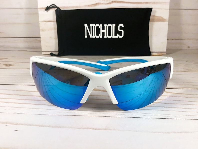 May include: White sports sunglasses with reflective blue lenses and bright blue temple tips. Behind the sunglasses is a black drawstring pouch featuring the white text NICHOLS printed in a bold, block-style font on the front surface.
