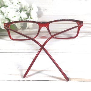 May include: Red rectangular eyeglasses with clear lenses. The frames have a textured top edge and thin temples. The glasses are set against a white wooden background with white flowers.