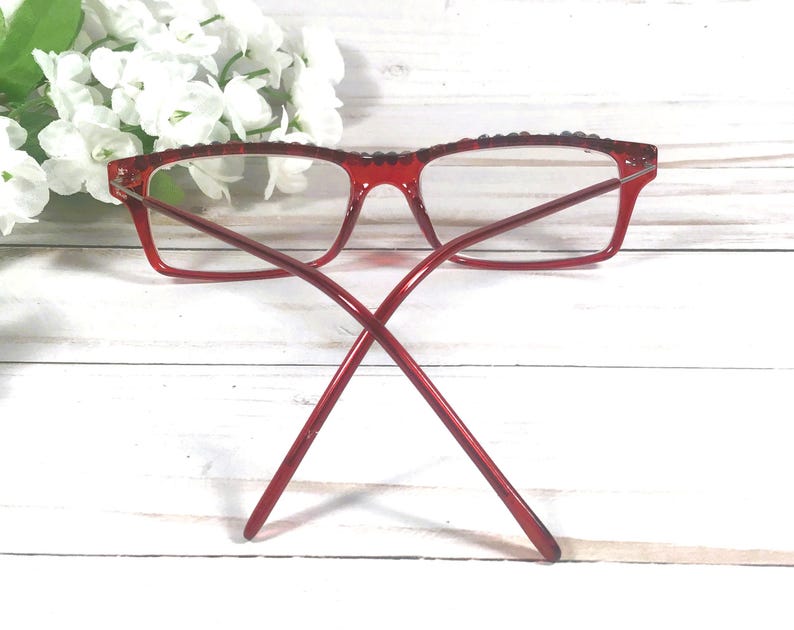 May include: Red rectangular eyeglasses with clear lenses. The frames have a textured top edge and thin temples. The glasses are set against a white wooden background with white flowers.
