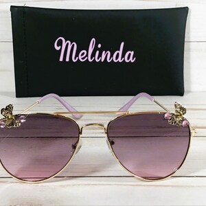 Op de afbeelding: Gouden aviator zonnebril met paarse glazen en vlinderaccenten op de pootjes. De zonnebril wordt geleverd met een zwarte etui met "Melinda" in roze letters.