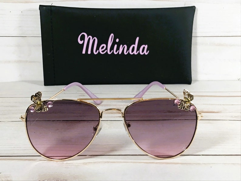 Op de afbeelding: Gouden aviator zonnebril met paarse glazen en vlinderaccenten op de pootjes. De zonnebril wordt geleverd met een zwarte etui met "Melinda" in roze letters.