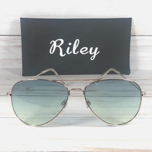 May include: Aviator sunglasses with a gold-coloured frame and gradient blue lenses. A black case with the name "Riley" in white script is behind the sunglasses. The sunglasses have a double bridge and a classic design.