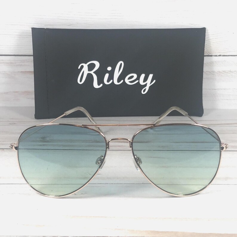 May include: Aviator sunglasses with a gold-coloured frame and gradient blue lenses. A black case with the name "Riley" in white script is behind the sunglasses. The sunglasses have a double bridge and a classic design.
