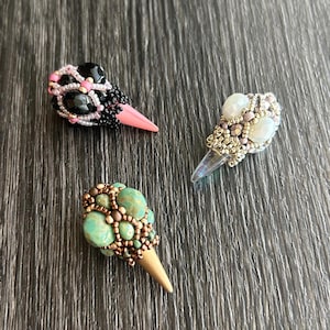 Könnte beinhalten: Drei perlenbesetzte, kegelförmige Ornamente mit unterschiedlich farbigen Perlen und einer spitzen Spitze. Die Ornamente befinden sich auf einer dunklen Holzoberfläche.