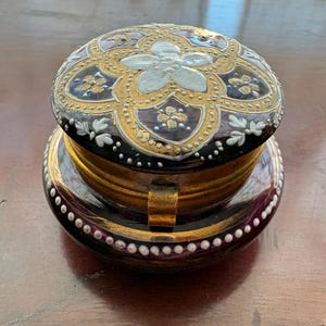 May include: An antique trinket box, predominantly purple with gold and white detailing. The lid features a floral pattern. The box is round with a hinged lid and a gold-coloured metal clasp. The box is likely made of glass or ceramic. The box is approximately 7 cm in diameter.