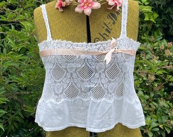 Puede incluir: Top blanco de encaje con una cinta y un lazo rosa. El top está en un maniquí con una guirnalda de flores rosas alrededor del cuello.