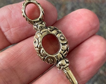 May include: A gold-toned watch key with an ornate, floral design. The key has a round head with a hole in the center and a long, thin shaft.