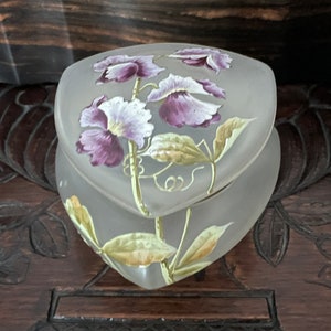 May include: A frosted glass trinket box in the shape of a heart with a floral design. The flowers are painted in shades of purple and green.
