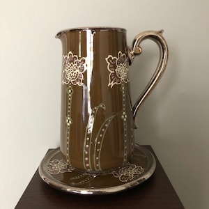 Puede incluir: Una jarra de cerámica marrón con asa plateada y diseño floral. La jarra está sobre un plato plateado a juego.