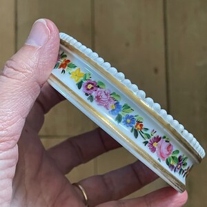 May include: A white porcelain trinket dish with a gold rim and a floral design. The dish has a beaded edge and is decorated with pink, yellow, blue, and red flowers.