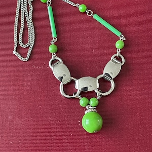 Puede incluir: Un collar de cadena de plata con un colgante de vidrio verde y cuentas de vidrio verde. El colgante es una cuenta redonda grande con una cuenta más pequeña en la parte superior. El collar está sobre un fondo rojo.