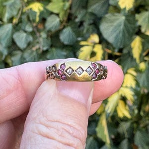 以下が含まれることがあります： 小さなダイヤモンドと赤い宝石が特徴のゴールドリング。リングは、側面に花のアクセントが付いた装飾的なデザインです。リングは緑の葉の背景に対して保持されています。