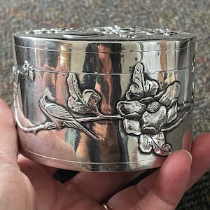 May include: A silver round trinket box with an embossed floral design featuring a bird and a flower. The box has a shiny finish.