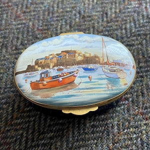 May include: A vintage oval-shaped trinket box with a painted scene of a harbor with boats and a town in the background. The box is made of metal with a gold-colored trim and a hinged lid.
