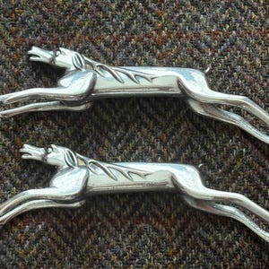 May include: Two silver metal greyhound figurines in a running pose. The dogs have detailed features and are set against a textured brown and green background. The figurines are likely decorative items.