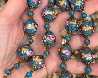 Könnte beinhalten: Eine Vintage-Halskette mit großen, runden, türkisfarbenen Perlen mit floralen Designs in Rosa, Gelb und Gold. Die Halskette hat kleinere türkisfarbene Perlen und einen goldfarbenen Verschluss. Die Perlen werden in einer Hand gehalten.