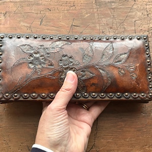 Puede incluir: Una caja rectangular de cuero marrón con un diseño floral. La caja presenta un patrón floral en relieve con flores y hojas. Los bordes están forrados con pequeños tachuelas metálicas redondas. La caja se sostiene en una mano.