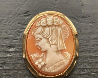 May include: Oval cameo brooch featuring a profile of a woman with detailed hair and clothing, set in a gold-colored frame. The cameo is a light orange color, and the frame has a polished finish. The brooch is a classic piece of jewelry.