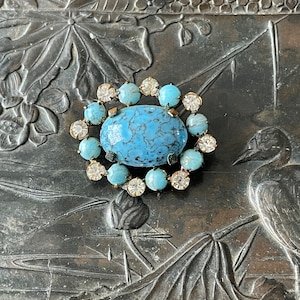May include: A turquoise blue oval cabochon stone surrounded by a ring of clear rhinestones set in a silver tone metal brooch.