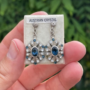 May include: A pair of silver dangle earrings with blue Austrian crystal stones. The earrings are on a white card with the words "Austrian Crystal" printed on it.